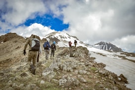 A group of hikers with backpacks is trekking up a rocky mountain slope, with patches of snow visible in the background. The sky is partly cloudy, creating a dynamic contrast with the bright blue color in between the clouds. The hikers are wearing outdoor gear appropriate for the cool weather.