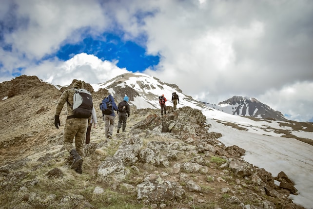 A group of hikers with backpacks is trekking up a rocky mountain slope, with patches of snow visible in the background. The sky is partly cloudy, creating a dynamic contrast with the bright blue color in between the clouds. The hikers are wearing outdoor gear appropriate for the cool weather.