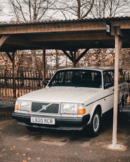 A classic white Volvo parked under a wooden carport with an autumnal background of leafless trees. The vehicle's registration plate is visible, and the ground is scattered with fallen leaves.