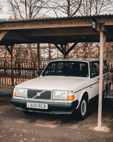 A classic white Volvo parked under a wooden carport with an autumnal background of leafless trees. The vehicle's registration plate is visible, and the ground is scattered with fallen leaves.