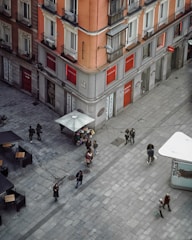 A street corner with a coral-colored building featuring multiple balconies and businesses on the ground floor. People walk along the wide, cobblestone pedestrian area. A vendor with a cart of flowers and a large umbrella offers a pop of color in the scene. Outdoor tables and chairs are visible along the street.