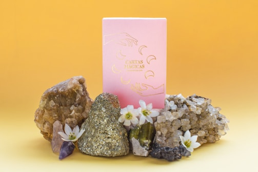 A pink box with gold illustrations stands surrounded by various crystals and small white flowers against a gradient yellow background. The display includes a pyrite stone, amethyst, and quartz clusters.