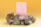 A pink box with gold illustrations stands surrounded by various crystals and small white flowers against a gradient yellow background. The display includes a pyrite stone, amethyst, and quartz clusters.