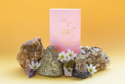 A pink box with gold illustrations stands surrounded by various crystals and small white flowers against a gradient yellow background. The display includes a pyrite stone, amethyst, and quartz clusters.