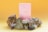 A pink box with gold illustrations stands surrounded by various crystals and small white flowers against a gradient yellow background. The display includes a pyrite stone, amethyst, and quartz clusters.