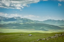 A rugged landscape featuring expansive green hills and distant snow-capped mountains under a partly cloudy blue sky. A solitary white SUV is parked on a grassy plain, contributing to a sense of isolation and exploration.