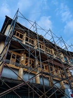 A multi-story building is under construction or renovation, surrounded by metal scaffolding. The structure features a series of wooden-framed windows and decorative railings. The sky above is mostly clear with patches of clouds.