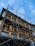 A multi-story building is under construction or renovation, surrounded by metal scaffolding. The structure features a series of wooden-framed windows and decorative railings. The sky above is mostly clear with patches of clouds.