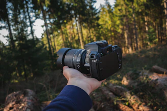 A candid shot of John Manuwal holding a digital camera outdoors, sunlight filtering through trees.
