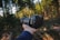 A person holds a DSLR camera in their right hand, positioned in a forest setting with tall trees and dappled sunlight filtering through the branches.