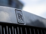 Close-up of a metallic surface with the Rolls-Royce emblem prominently displayed. The emblem consists of two overlapping 'R' letters with the words 'ROLLS' and 'ROYCE' above and below the letters. The background is softly blurred, giving a focus to the emblem.