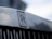 Close-up of a metallic surface with the Rolls-Royce emblem prominently displayed. The emblem consists of two overlapping 'R' letters with the words 'ROLLS' and 'ROYCE' above and below the letters. The background is softly blurred, giving a focus to the emblem.
