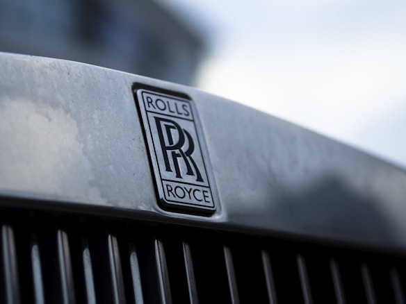 Close-up of a metallic surface with the Rolls-Royce emblem prominently displayed. The emblem consists of two overlapping 'R' letters with the words 'ROLLS' and 'ROYCE' above and below the letters. The background is softly blurred, giving a focus to the emblem.