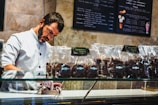 A man in a white button-up uniform behind a glass display case is carefully packaging items, wearing gloves. Several transparent bags filled with chocolate or cookies are lined up on the counter. The background features menu boards with various desserts listed.