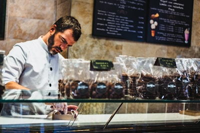 A man in a white button-up uniform behind a glass display case is carefully packaging items, wearing gloves. Several transparent bags filled with chocolate or cookies are lined up on the counter. The background features menu boards with various desserts listed.