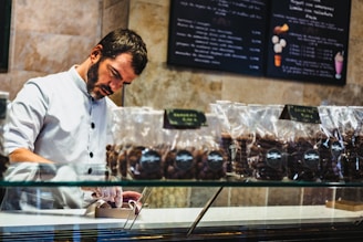 A man in a white button-up uniform behind a glass display case is carefully packaging items, wearing gloves. Several transparent bags filled with chocolate or cookies are lined up on the counter. The background features menu boards with various desserts listed.