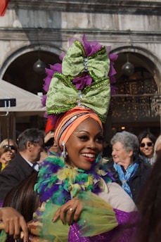 A woman is joyfully participating in a festival, wearing a colorful outfit with a large, elaborate headpiece featuring purple and green bows. She is smiling brightly, surrounded by a crowd in a festive atmosphere.