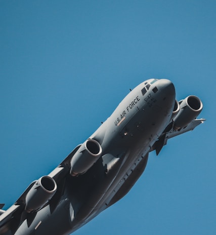 A U.S. Air Force military aircraft captured from a low angle against a clear blue sky. The aircraft appears to be a large cargo plane, prominently displaying its engines and part of its fuselage.