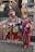 Two individuals dressed in vibrant, colorful costumes with elaborate masks are standing in a public space. Their outfits are adorned with various ornaments and patterns, including patchwork designs and sequins. Both are holding ornate staffs. Behind them, a group of people are walking on a bridge in an urban setting with old architecture.