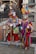 Two individuals dressed in vibrant, colorful costumes with elaborate masks are standing in a public space. Their outfits are adorned with various ornaments and patterns, including patchwork designs and sequins. Both are holding ornate staffs. Behind them, a group of people are walking on a bridge in an urban setting with old architecture.