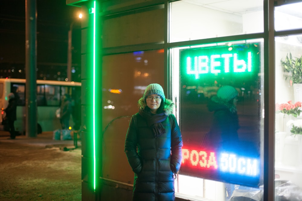 woman wearing black bubble coat and gray knit cap standing beside building with LED signage at night