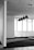 A modern office space featuring several large windows allowing ample natural light. The ceiling has a grid-like pattern with multiple hanging lights. The floor is carpeted and the walls are mostly blank, contributing to a minimalist aesthetic.