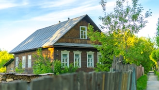 A rustic wooden house with a sloped metal roof surrounded by lush greenery. The building features several white-framed windows and is bordered by a wooden fence. The scene is illuminated by bright, natural sunlight creating a serene and peaceful rural setting.