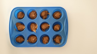 A blue silicone muffin tray with twelve cavities, each filled with a dollop of brown dough, is placed on a white surface.