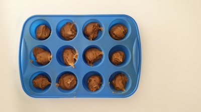 A blue silicone muffin tray with twelve cavities, each filled with a dollop of brown dough, is placed on a white surface.
