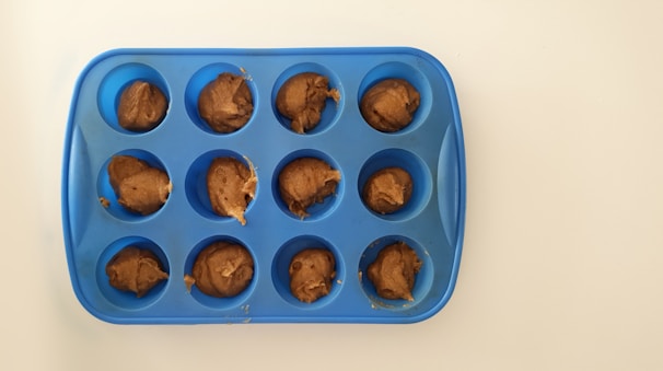 A blue silicone muffin tray with twelve cavities, each filled with a dollop of brown dough, is placed on a white surface.