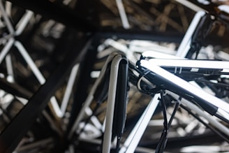 A complex network of interwoven metal pipes and tubes illuminated by white neon lights creates an abstract, modern art installation. The geometric arrangement produces a visually striking pattern that plays with light and shadow.