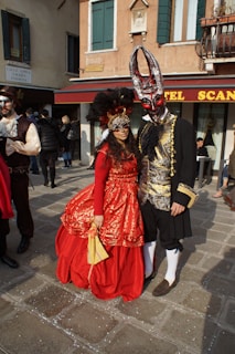 Two people dressed in elaborate, ornate costumes from a historical or fictional setting are standing in a paved street. The person on the left wears a red dress with intricate gold detailing, a matching mask, and a large headdress with black feathers. The person on the right wears a black outfit with gold embroidery, white stockings, and a mask with long horns. They are standing in front of a building with red awnings and teal shutters, creating a backdrop that suggests a festive or theatrical event.