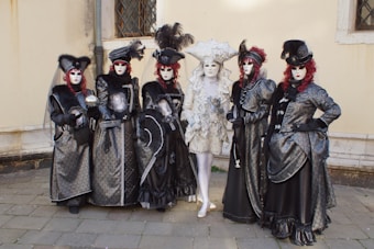 A group of six individuals dressed in elaborate and ornate costumes, featuring rich fabrics and intricate details. They are wearing masks and extravagant hats, with one individual in the center wearing a white and silver costume, while the others are in black and dark silver attire with red wigs.