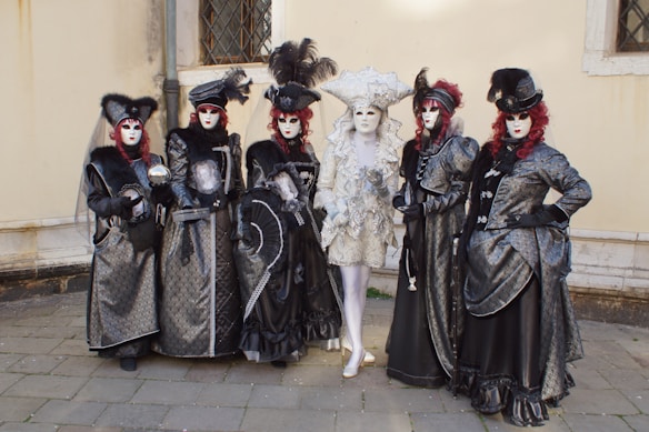 A group of six individuals dressed in elaborate and ornate costumes, featuring rich fabrics and intricate details. They are wearing masks and extravagant hats, with one individual in the center wearing a white and silver costume, while the others are in black and dark silver attire with red wigs.
