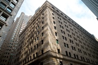 A large, multi-story stone building with numerous windows, surrounded by other tall, urban architectural structures. The foreground building features decorative arches and stonework, creating a sense of grandeur and solidity.