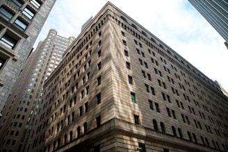 A large, multi-story stone building with numerous windows, surrounded by other tall, urban architectural structures. The foreground building features decorative arches and stonework, creating a sense of grandeur and solidity.
