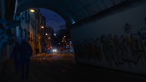 A dimly lit urban street scene during nighttime. Buildings line the path, adorned with bright street lights. The street is bordered by a concrete wall covered in colorful graffiti. Two people are walking towards a tunnel, while a tram can be seen approaching down the tracks.