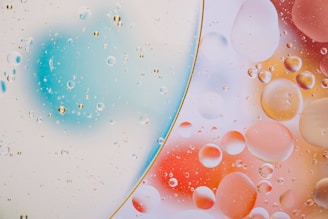 A close-up view of oil droplets suspended in water on a glass surface, forming an abstract pattern. The droplets are of various sizes and are scattered throughout, creating a visually intriguing texture against a colorful backdrop of blurred blue, orange, and pink hues. The scene is divided by a curved line, suggesting a play with surface tension and refraction.