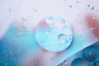 A multitude of transparent bubbles are suspended in a liquid, set against a background blending hues of blue, pink, and white. The bubbles vary in size and are overlapping in some areas, creating an effect of depth and dimensionality.