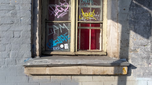 A weathered brick wall features a vintage-style window adorned with colorful graffiti. Various shades of vibrant spray paint create intricate designs and tagging on the window panes and frame. A ledge beneath the window appears worn, adding to the urban, street-art feel of the scene.