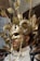 A person is wearing an elaborate costume featuring a gold mask with intricate designs and large curved horns. The attire includes gold glitter, feathers, chains, and decorative elements such as leaves. The overall style is theatrical and extravagant.