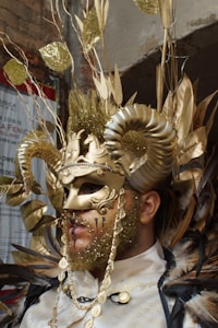 A person is wearing an elaborate costume featuring a gold mask with intricate designs and large curved horns. The attire includes gold glitter, feathers, chains, and decorative elements such as leaves. The overall style is theatrical and extravagant.