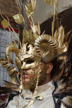 A person is wearing an elaborate costume featuring a gold mask with intricate designs and large curved horns. The attire includes gold glitter, feathers, chains, and decorative elements such as leaves. The overall style is theatrical and extravagant.
