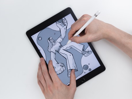 A hand uses a stylus to draw on a tablet, depicting a cartoon character in a casual pose. The digital drawing illustrates a laid-back character with long hair wearing headphones, large boots, and baggy clothing, surrounded by abstract shapes.