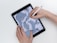 A hand uses a stylus to draw on a tablet, depicting a cartoon character in a casual pose. The digital drawing illustrates a laid-back character with long hair wearing headphones, large boots, and baggy clothing, surrounded by abstract shapes.