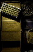 A tray of ammunition cartridges is placed on top of two boxes labeled 'Winchester, 50 Cartridges, Caliber 9mm, Full Metal Jacket, Service Grade.' Beside the boxes is a round plastic container filled with additional loose cartridges. The scene is dimly lit with a shadowy ambiance.