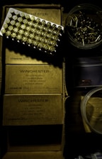 A tray of ammunition cartridges is placed on top of two boxes labeled 'Winchester, 50 Cartridges, Caliber 9mm, Full Metal Jacket, Service Grade.' Beside the boxes is a round plastic container filled with additional loose cartridges. The scene is dimly lit with a shadowy ambiance.