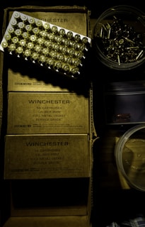 A tray of ammunition cartridges is placed on top of two boxes labeled 'Winchester, 50 Cartridges, Caliber 9mm, Full Metal Jacket, Service Grade.' Beside the boxes is a round plastic container filled with additional loose cartridges. The scene is dimly lit with a shadowy ambiance.