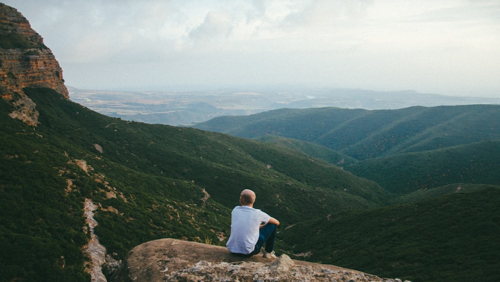 pessoa sentada à beira de um penhasco sobre montanhas durante o dia