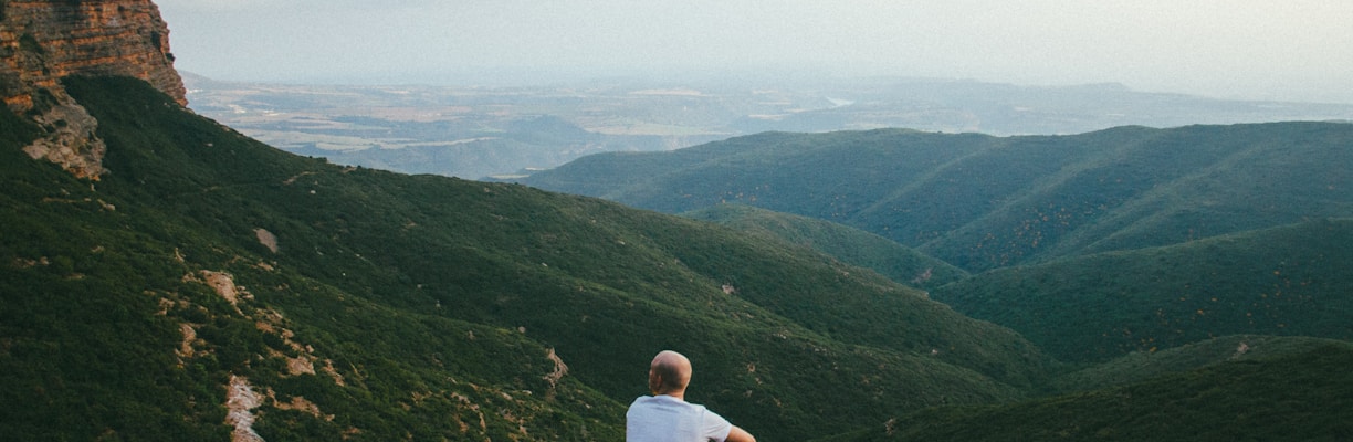 person sitting on the edge of a cliff over looking mountains during daytime